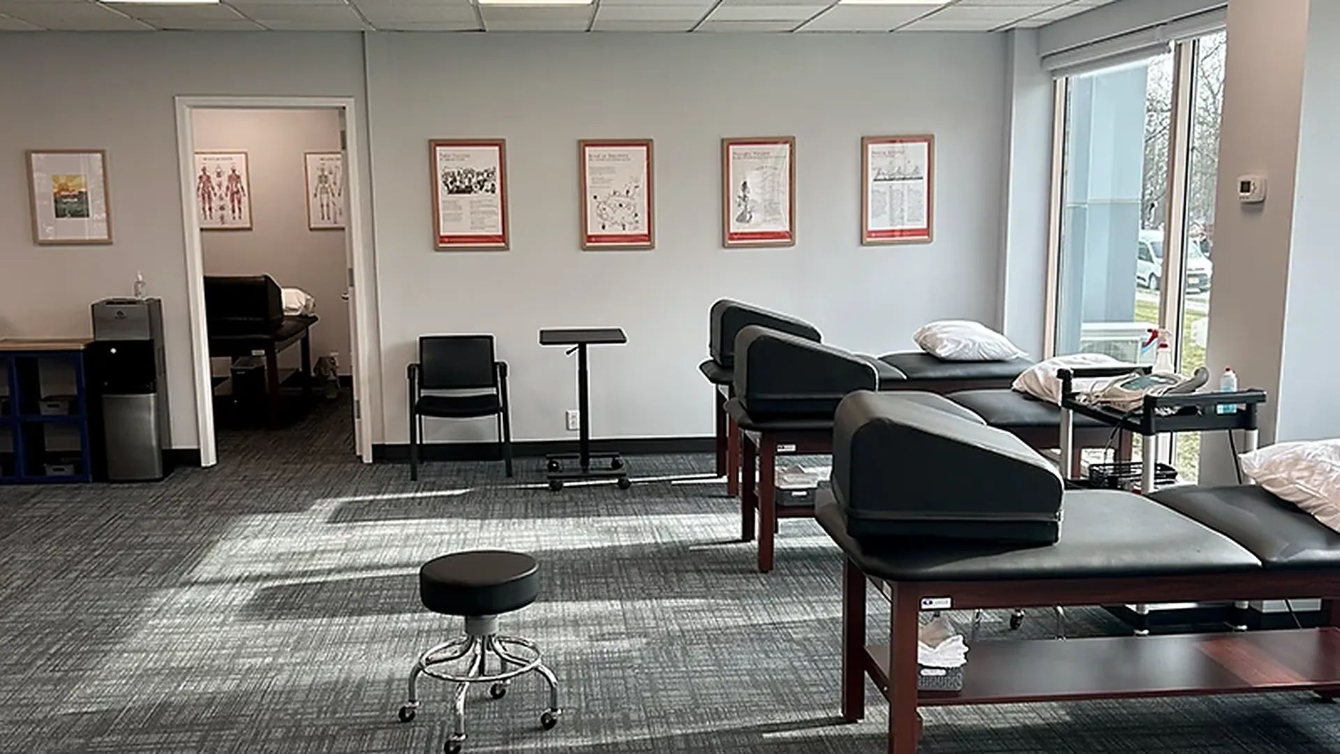 Treatment room at Carroll Physical Therapy with framed anatomy charts on the wall, padded treatment tables with wedge bolsters, and a rolling stool