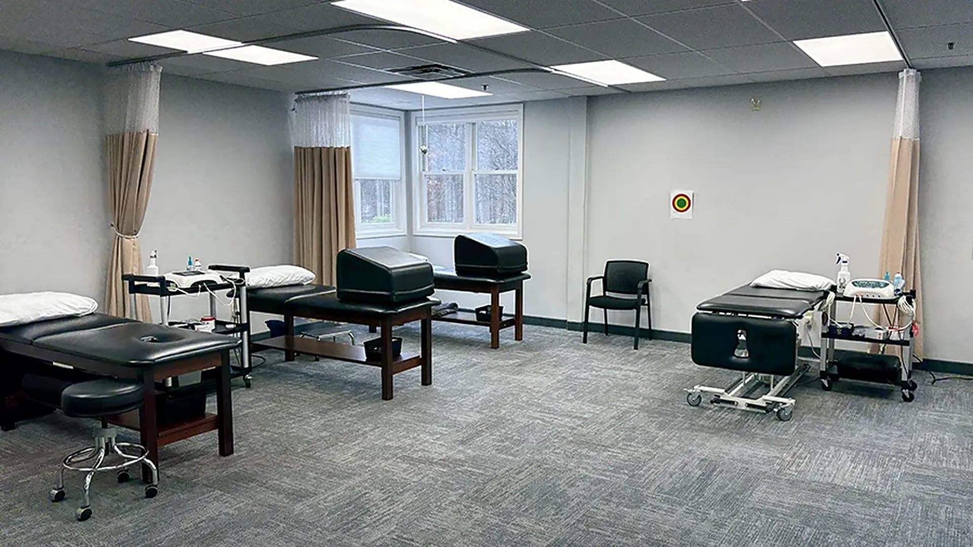 A treatment bay at a Carroll Physical Therapy clinic, with treatment tables and bolsters, privacy curtains, a hydraulic hi-lo table, a modality cart, and a bay window