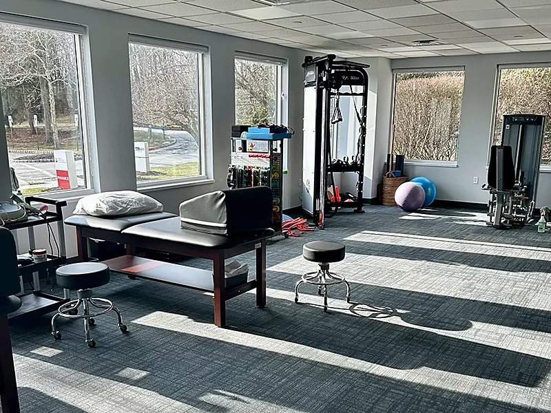 Treatment tables, stools, a cable pulley tower, and exercise balls on the sunlit rehab floor at the Mountain Lakes clinic