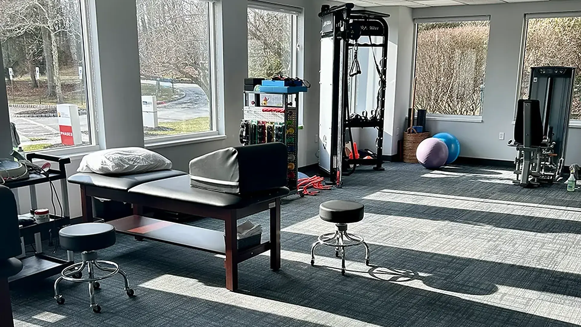 The exercise floor at Carroll Physical Therapy in Mountain Lakes, NJ, with a treatment table by the windows, a cable machine, resistance bands and stability balls