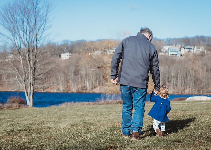 An older man walking hand in hand with a small child on the grass beside a lake in Morris County, NJ